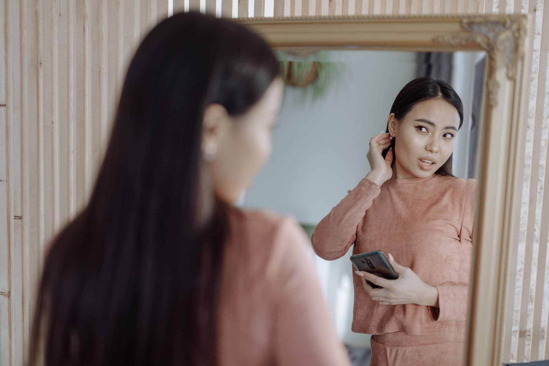 Asian woman reflects in mirror with smartphone wearing loungewear indoors.